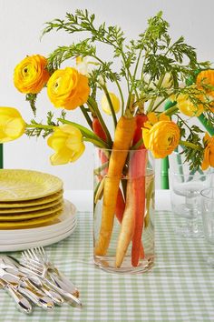 carrots and yellow flowers in a glass vase on a table with place settings for dinner