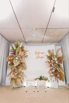 an indoor wedding setup with flowers and greenery on the wall, along with two white chairs