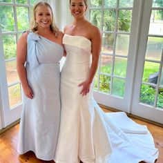 two women standing next to each other in front of large windows wearing white gowns