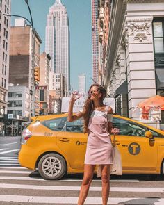 a woman standing in front of a taxi cab drinking from a paper bag while wearing sunglasses