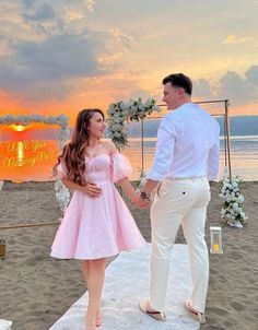 a man and woman standing on top of a sandy beach next to the ocean at sunset