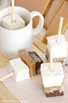 marshmallows and chocolate are on sticks in front of a cup of coffee