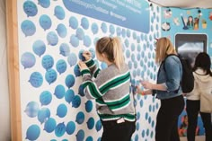 two women are painting a wall with blue circles on it and another woman is standing next to the wall