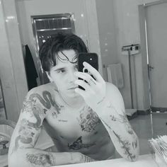 a man with tattoos taking a selfie in front of his phone while sitting at a bathroom sink