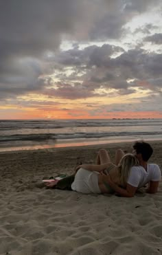 a man and woman sitting on top of a sandy beach next to the ocean at sunset