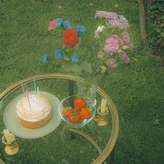 a glass table with a cake and flowers on it in the middle of a grassy area