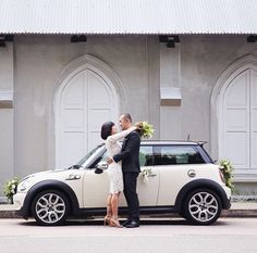 a bride and groom kissing in front of a white mini cooper wedding car with flowers on the hood