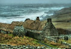 an old stone house with thatched roof next to the ocean on a cloudy day