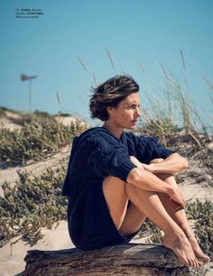 a woman sitting on top of a log in the sand with her legs crossed and looking off into the distance