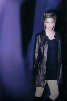 a woman standing in front of a blue background wearing a black dress and leather jacket