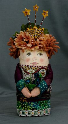 a small doll with red hair and flowers on it's head, sitting in front of a gray background