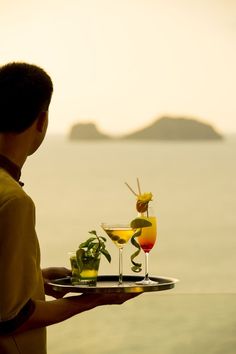 a man is holding a tray with two drinks on it and the ocean in the background