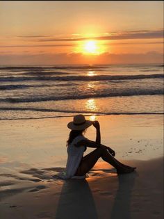 a woman sitting on top of a sandy beach next to the ocean at sun set