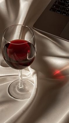 a glass of red wine sitting next to a laptop on a white satin tablecloth