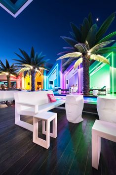 an outdoor lounge area with white benches and palm trees in front of the pool at night