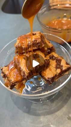 someone pouring caramel sauce on some brownies in a glass bowl with other dishes behind them