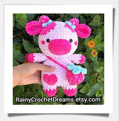 a hand holding a pink and white crocheted teddy bear in front of green leaves