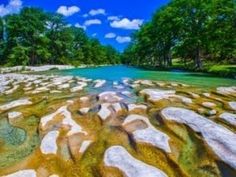 a river with rocks and trees in the water near some green grass, blue sky and white clouds