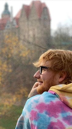 a man with glasses and a tie dye hoodie looks off into the distance as he talks on his cell phone