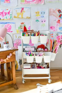 a white cart with lots of craft supplies on it in front of a wall full of children's artwork