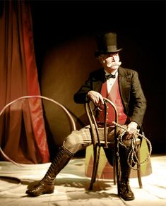 a man sitting in a chair wearing a top hat and holding a whip with one hand