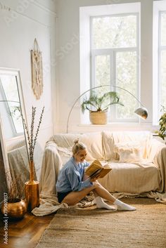 a woman sitting on the floor reading a book in front of a window with potted plants