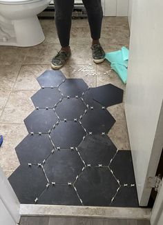 a person standing in front of a toilet on a bathroom floor with black hexagonal tiles