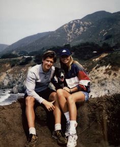 a man and woman sitting next to each other on top of a hill near the ocean