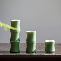 three green vases sitting next to each other on top of a wooden table with a plant in the background