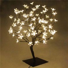 a lighted tree with white flowers is shown on a black base and stands against a gray background