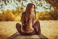 a woman sitting on top of a wooden bench next to a tree in a field