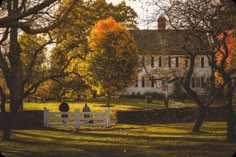 two people sitting on a bench in front of a large house with trees around them