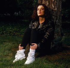 a woman sitting in the grass next to a tree with her hands on her knees