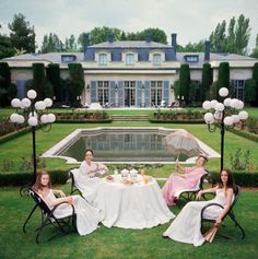 three women sitting at a table in front of a large house with a pool and fountain