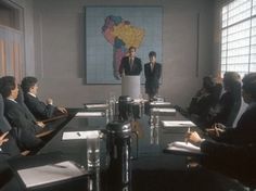 a group of people sitting around a conference room table in front of a man giving a speech