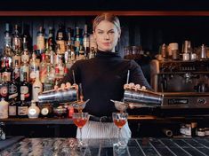 a woman holding two cocktail glasses in front of a bar filled with bottles and liquors