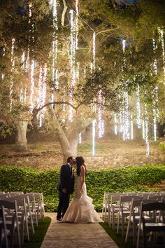 a bride and groom standing under a tree with lights hanging from it's branches