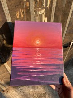 a person holding up a painting with the sun setting in the sky over water on it