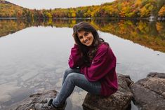 a woman sitting on top of a rock next to a lake surrounded by fall foliage