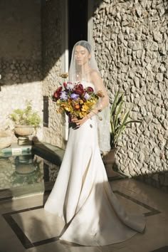 a woman in a wedding dress holding a bouquet of flowers next to a stone wall