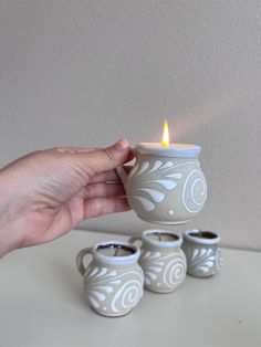 a hand holding a lit candle in front of four ceramic mugs on a table