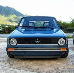 the front end of a blue car parked in a parking lot with mountains in the background