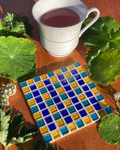 a cup of tea sits next to some green leaves and a mosaic tile coaster on a wooden table