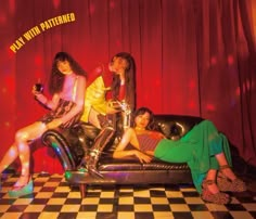 three women sitting on top of a couch in front of a checkered floor and red curtain