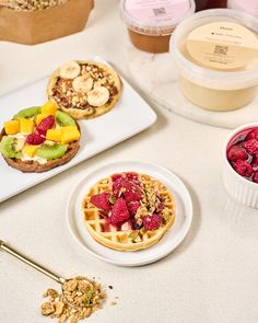 several different types of waffles and fruit on a white table top with containers of yogurt in the background