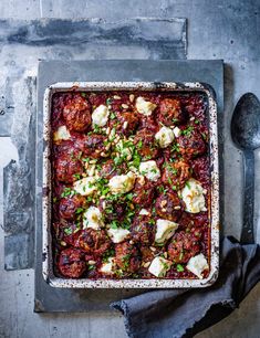 a pan filled with meatballs and cheese on top of a table next to a spoon