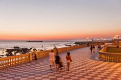 several people walking on a walkway next to the ocean at sunset or dawn with lights in the distance