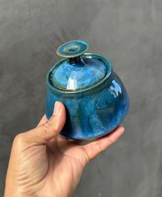 a hand holding a small blue pot with a lid on it's side, in front of a gray background