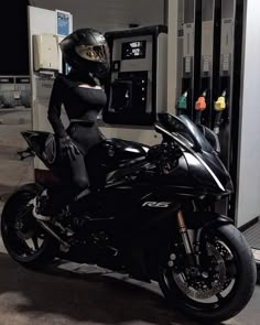 a woman in black is sitting on a motorcycle at a gas station and posing for the camera