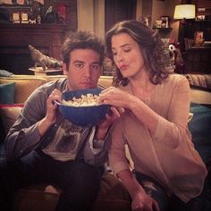 a man and woman sitting on a couch eating popcorn from a blue bowl while looking at the camera